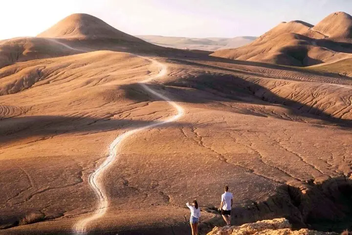 Two people on a ridge above a track winding through bare, rolling desert hills