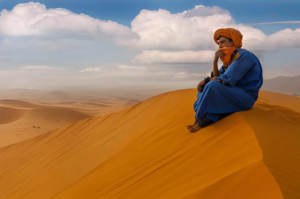 A man in a blue robe and orange turban on the crest of a sand dune