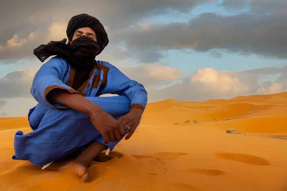 A man in a blue robe and black turban sitting on orange sand dunes