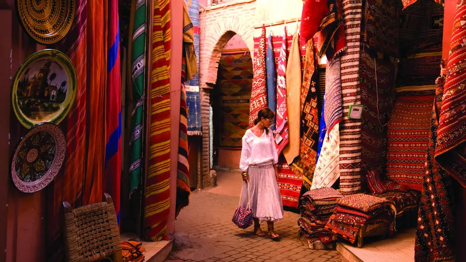 A woman walking a narrow souk lane hung with rugs and textiles