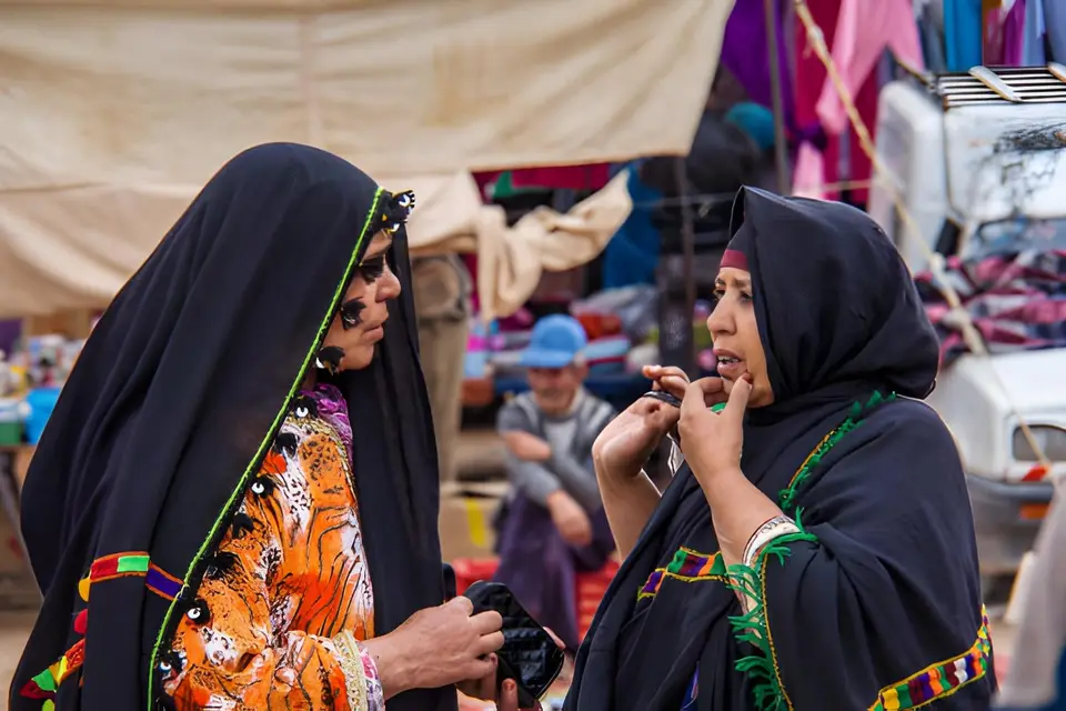 Two women in dark, patterned wraps talking in a busy market