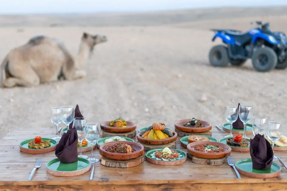Moroccan dishes laid out on a table in the desert, with a camel and a quad bike behind