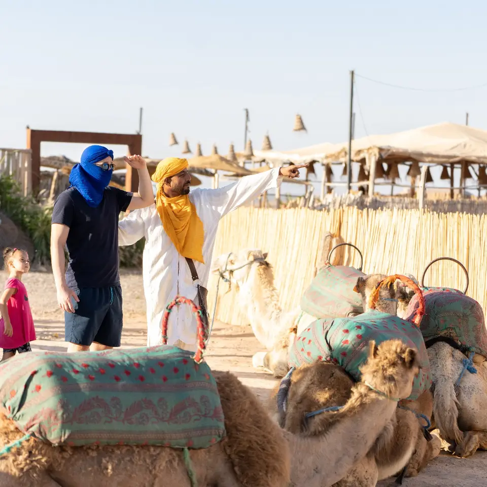 A guest in a blue headscarf and a camel handler beside camels at a desert camp